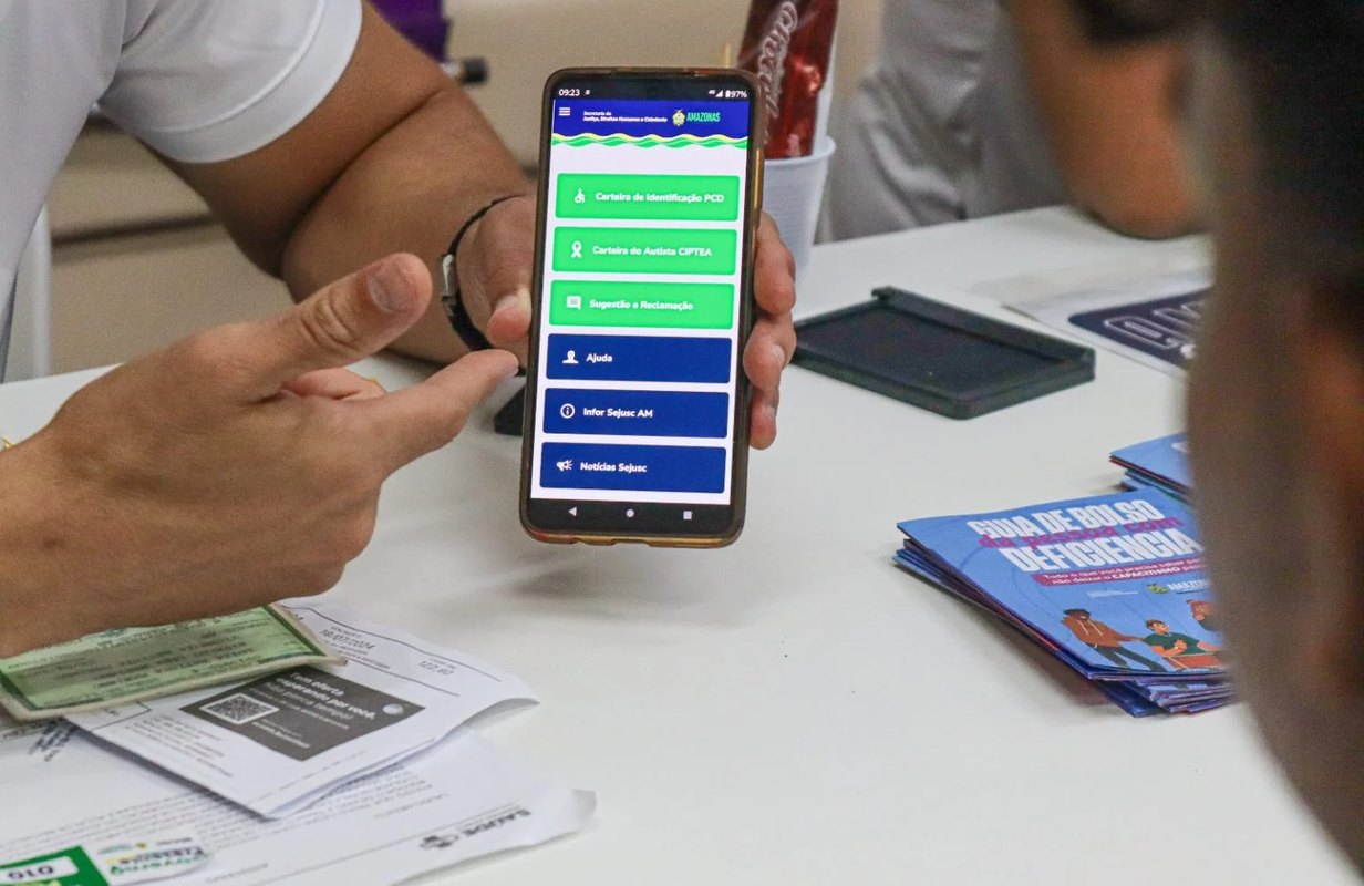 A SEJUSC officer showing the PCD Digital app on a phone at a service desk, with ID documents and leaflets on the table